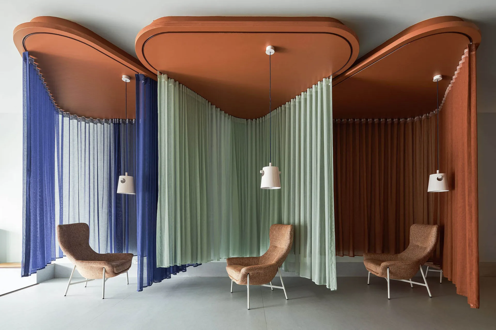 A modern living room with three wavy, curved textile dividers creating individual seating areas. Each divider is suspended fr