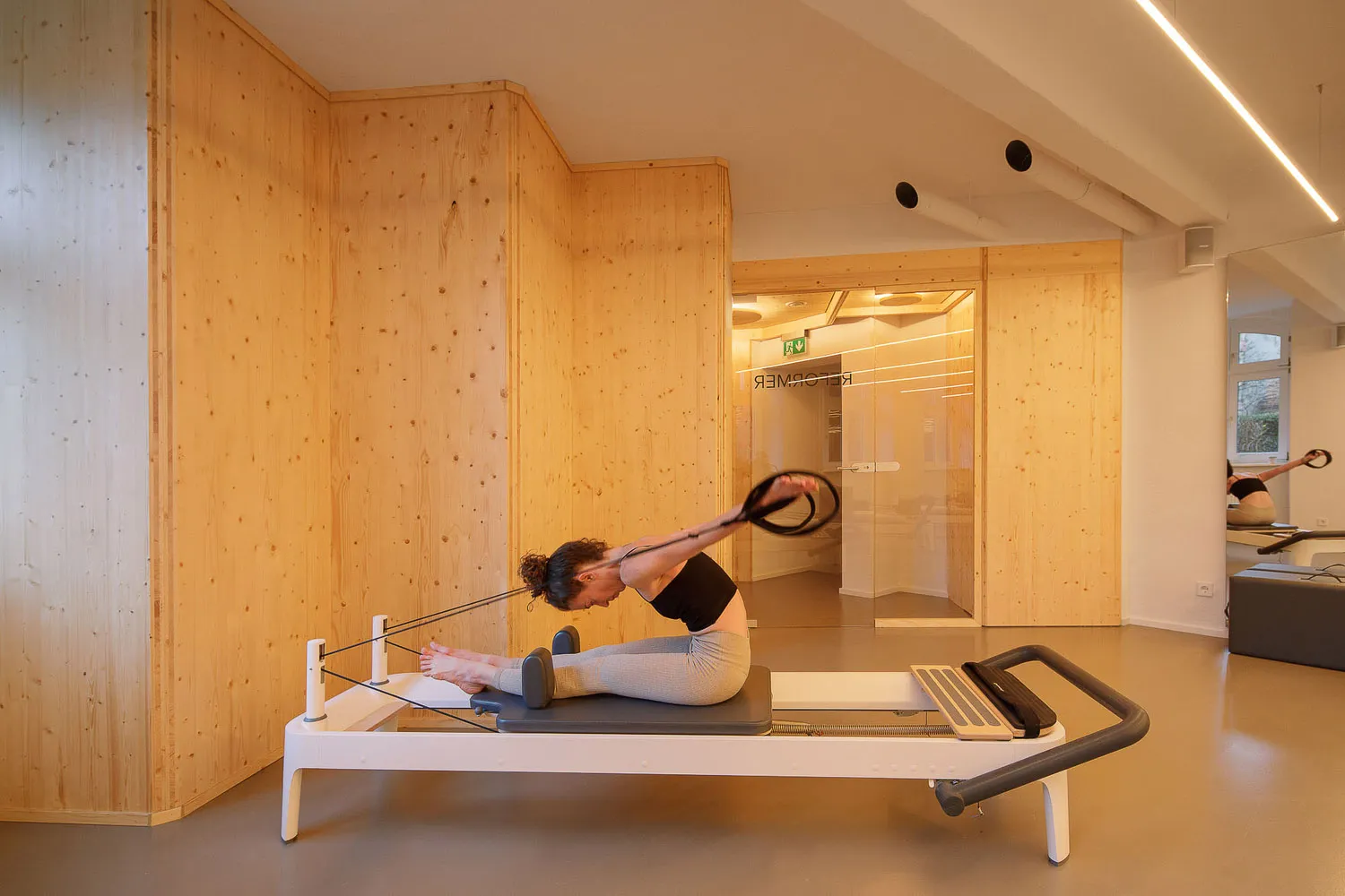 A woman performing yoga on a modern wooden fitness center platform in a spacious, minimalist room with large glass doors.