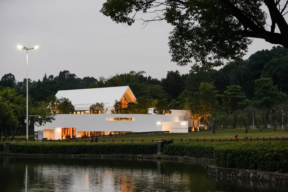 A serene outdoor view of a modern white concrete building with warm indoor lighting and a reflective pond.