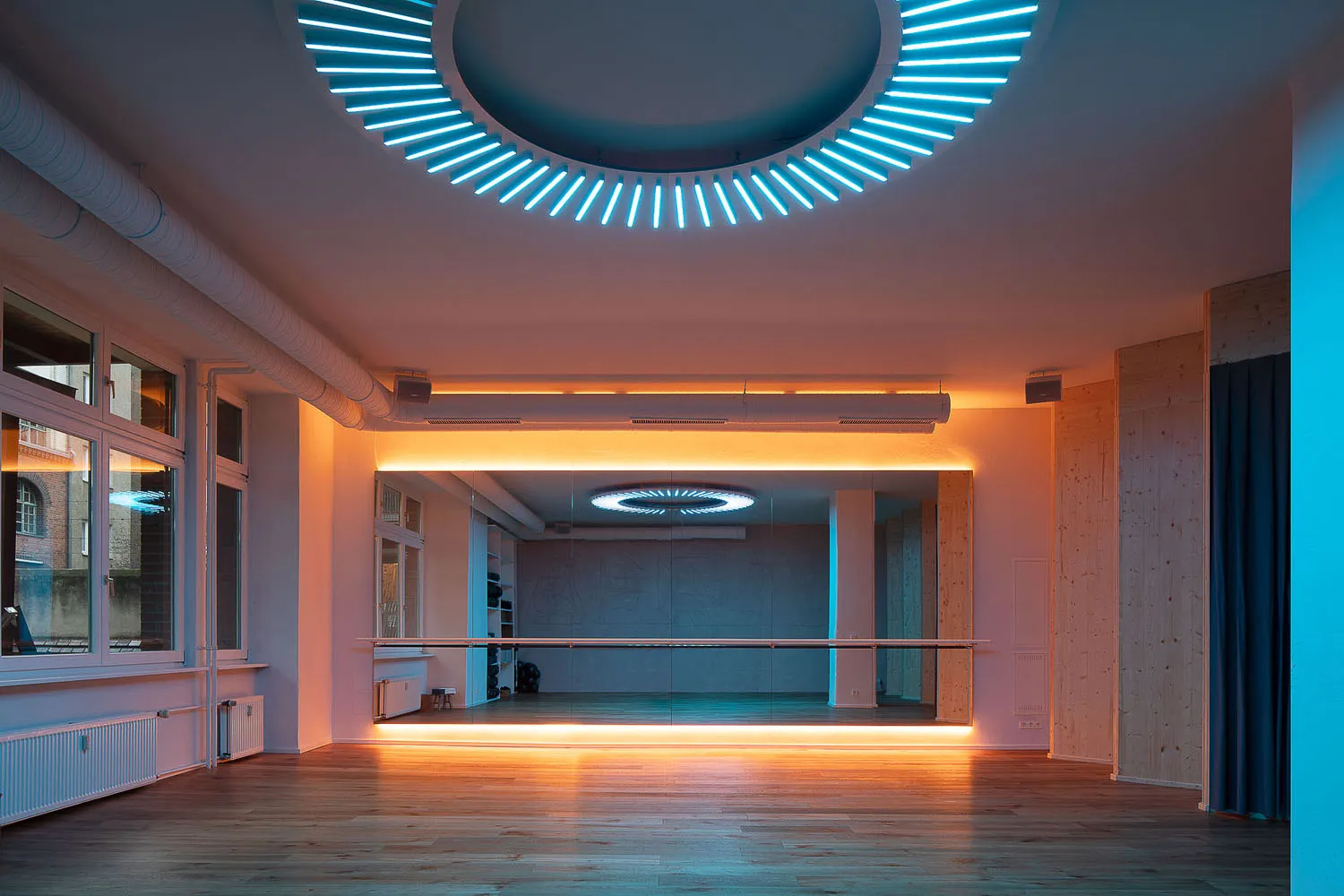 A spacious, modern dance studio with hardwood floors and glass walls, illuminated by neon lights and soft ambient lighting.