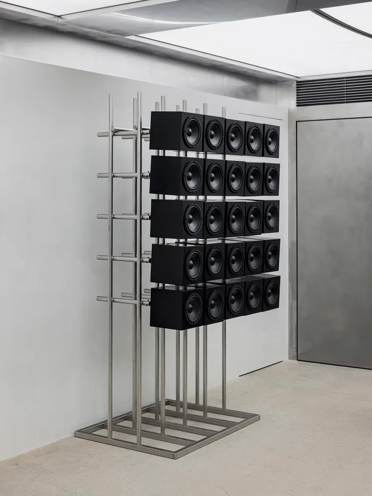 A minimalist audio system display made of metal featuring multiple speakers arranged in a grid on a metallic stand in a dimly