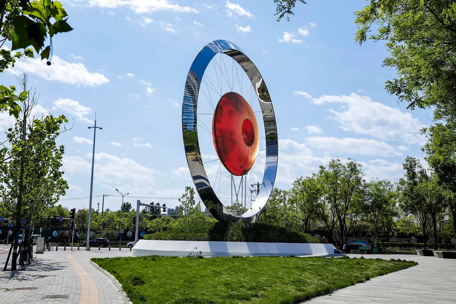 A large metallic circular sculpture with a red glass center, set in a park-like setting with greenery and pathways.