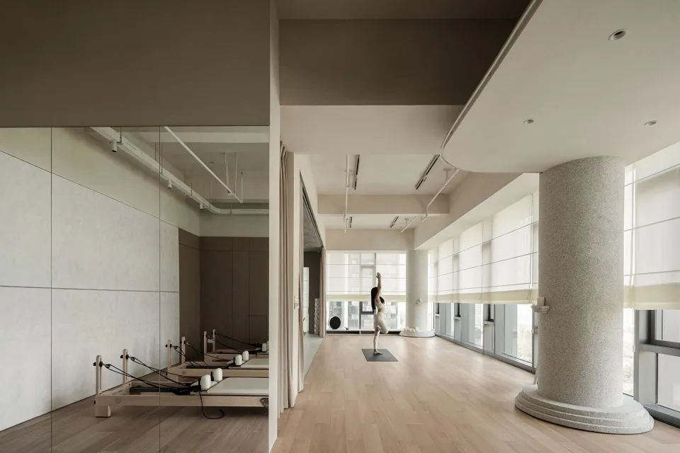 A modern fitness studio with wooden flooring and concrete walls, featuring Pilates reformers and a woman practicing yoga.