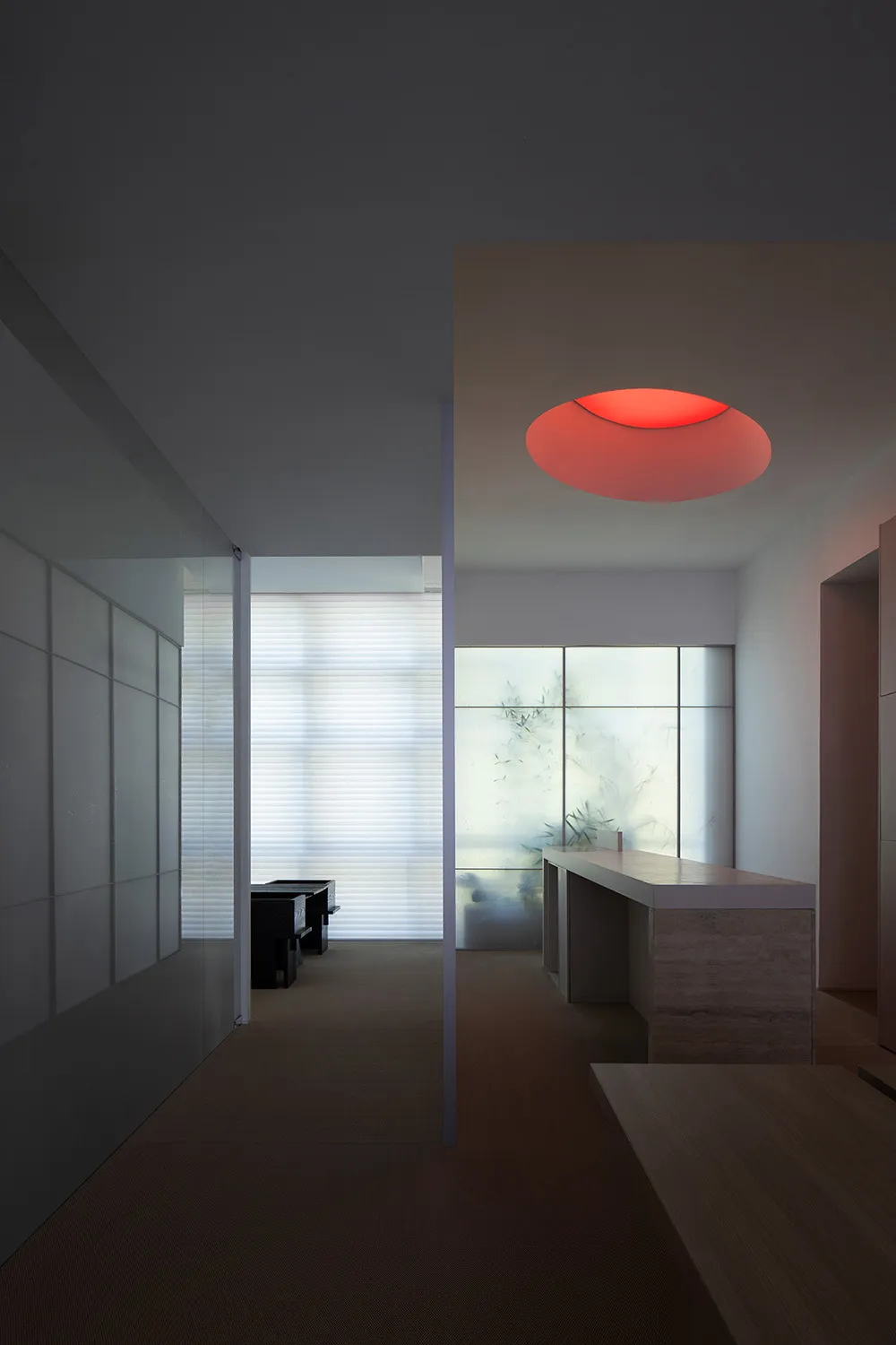 A modern minimalist kitchen with white surfaces, a red circular light fixture, and a view to a frosted glass window.