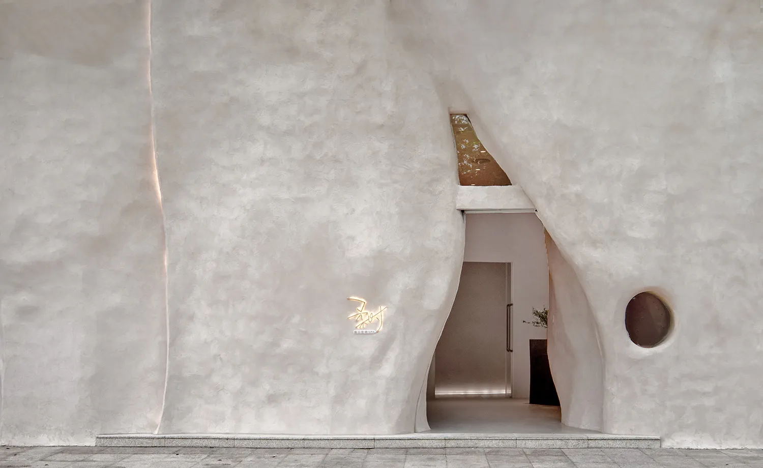 A modern architectural design featuring a textured concrete exterior wall with a circular window and a curved entryway.