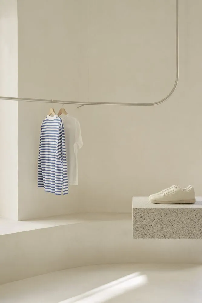 A minimalist white interior features a concrete bench with a pair of sneakers placed on it, a white tee, a blue and white str
