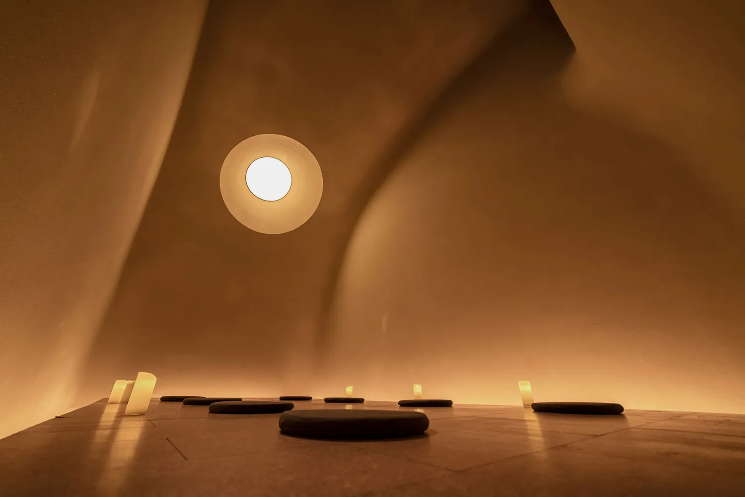 A minimalist, concrete-walled room with a circular skylight and lit candles arranged on the floor.