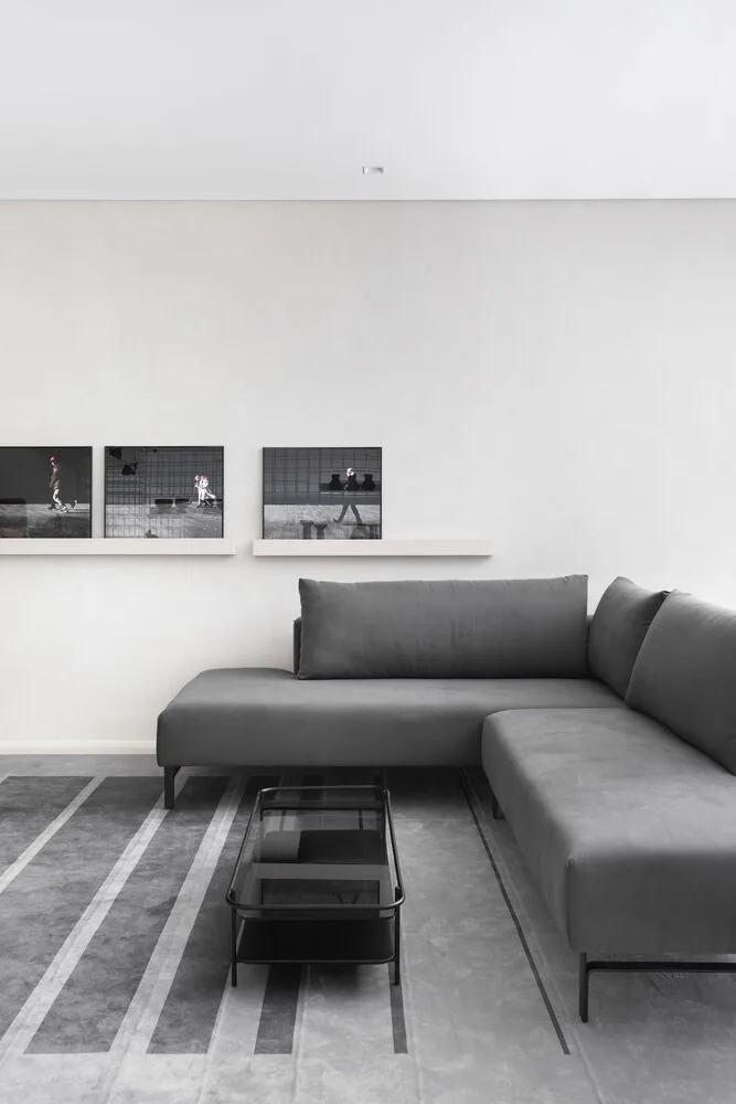 A minimalist living room featuring a gray sectional sofa on a patterned concrete floor, with a glass coffee table and framed