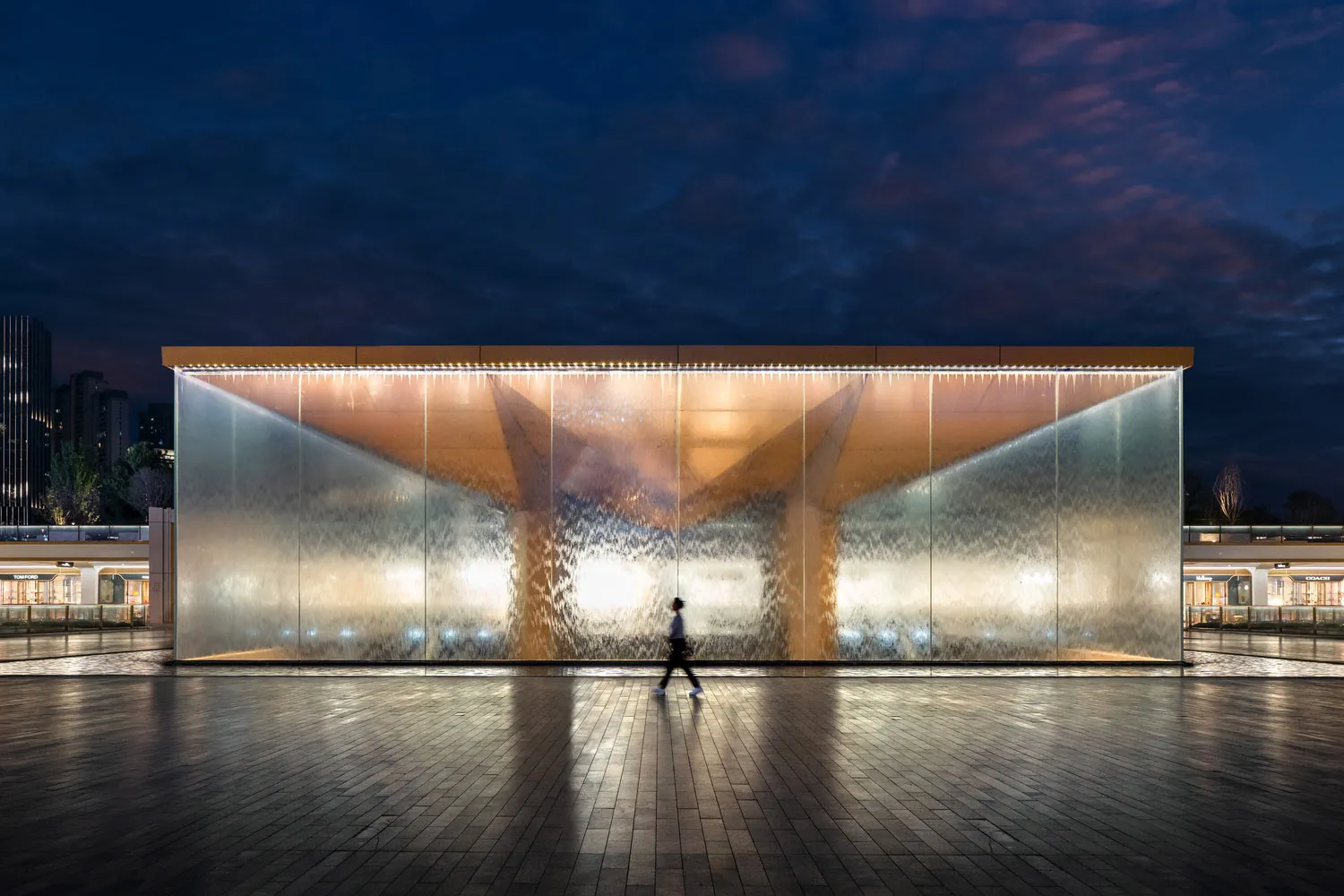 A modern public space featuring a glass water feature illuminated at night with a person walking in front.