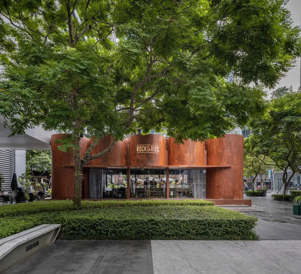 A modern restaurant exterior featuring a metal corrugated facade under a lush tree canopy.