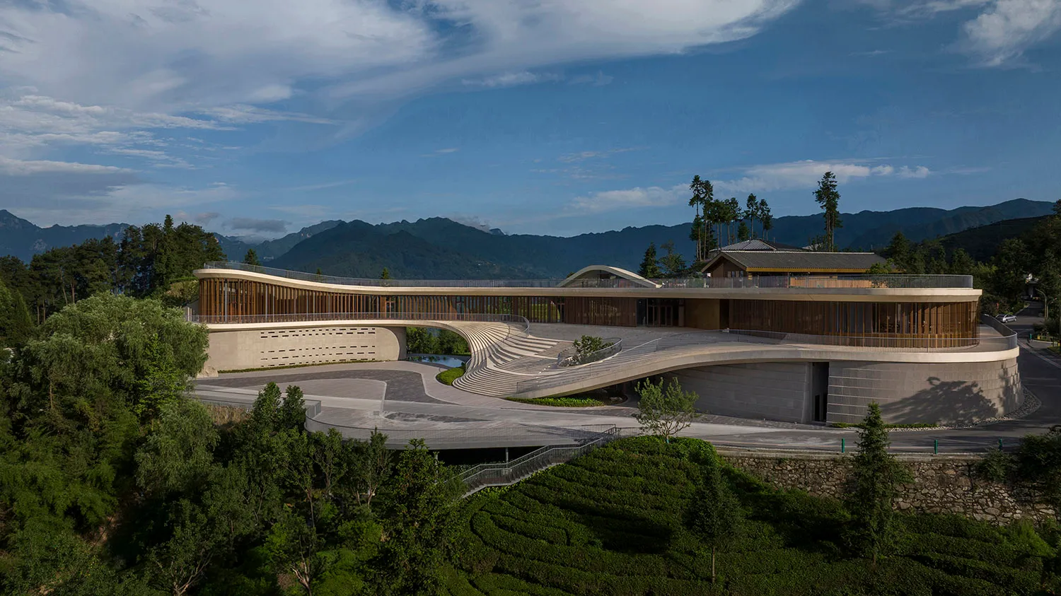 A modern hotel with concrete and glass exterior, surrounded by greenery and mountains.