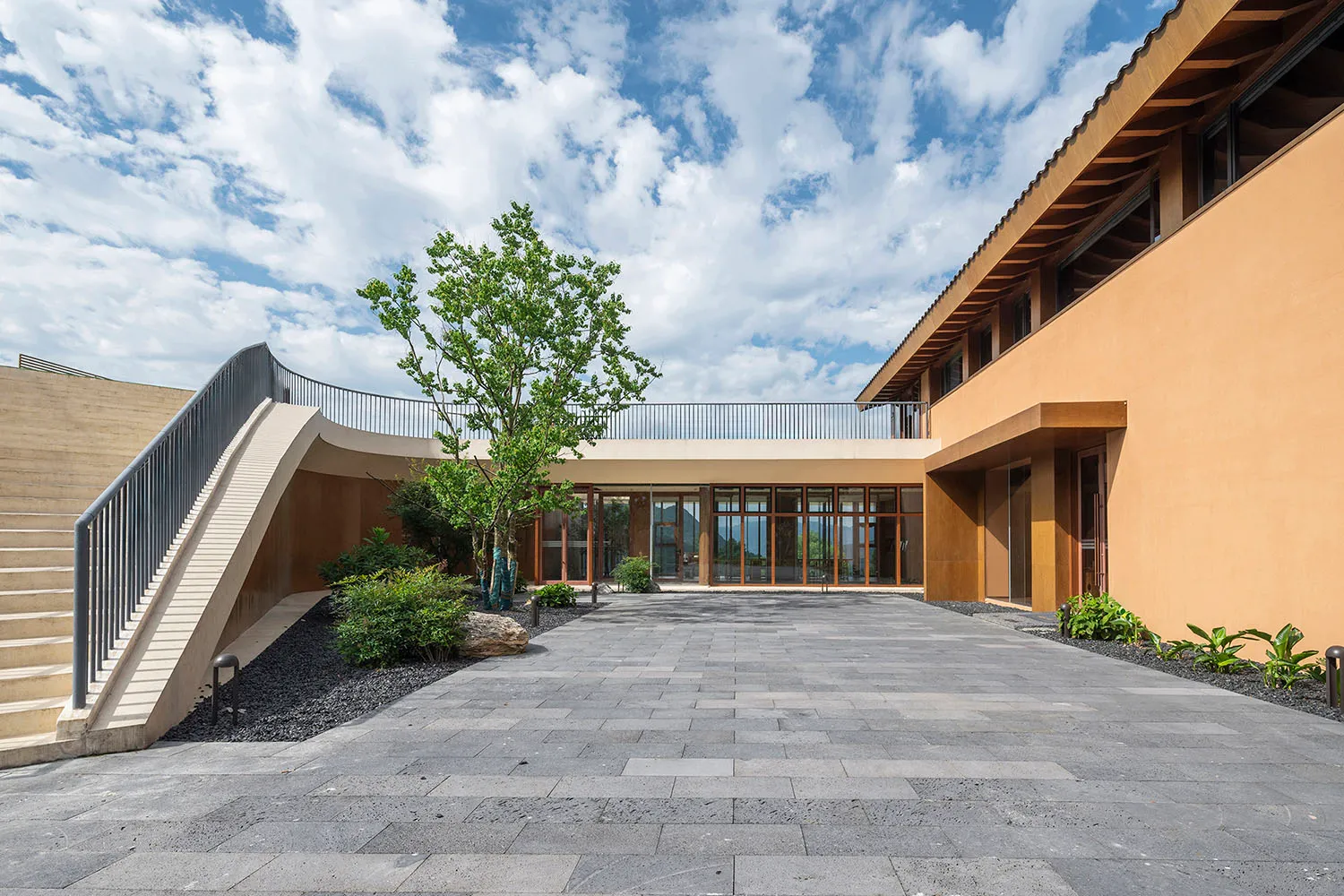 Modern office building exterior with concrete and glass materials, featuring a curved staircase and a central tree.
