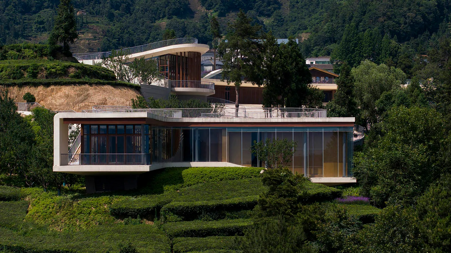 Modern residential house with concrete exterior, surrounded by greenery and hills in the background.
