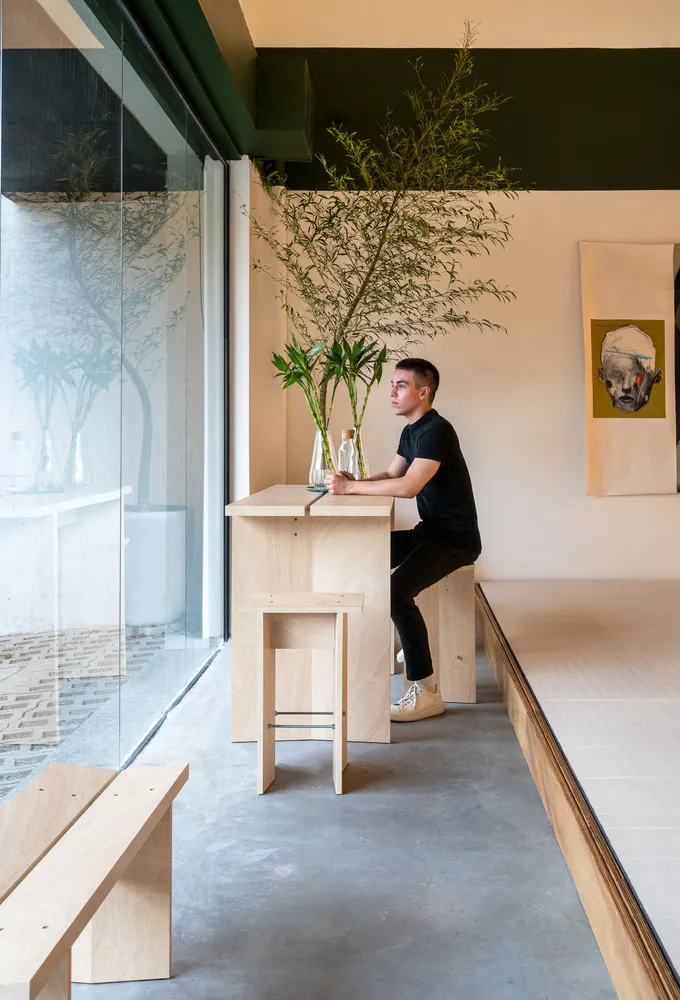 Person sitting at a minimalist restaurant interior with wood and concrete elements, plants, and a portrait artwork.