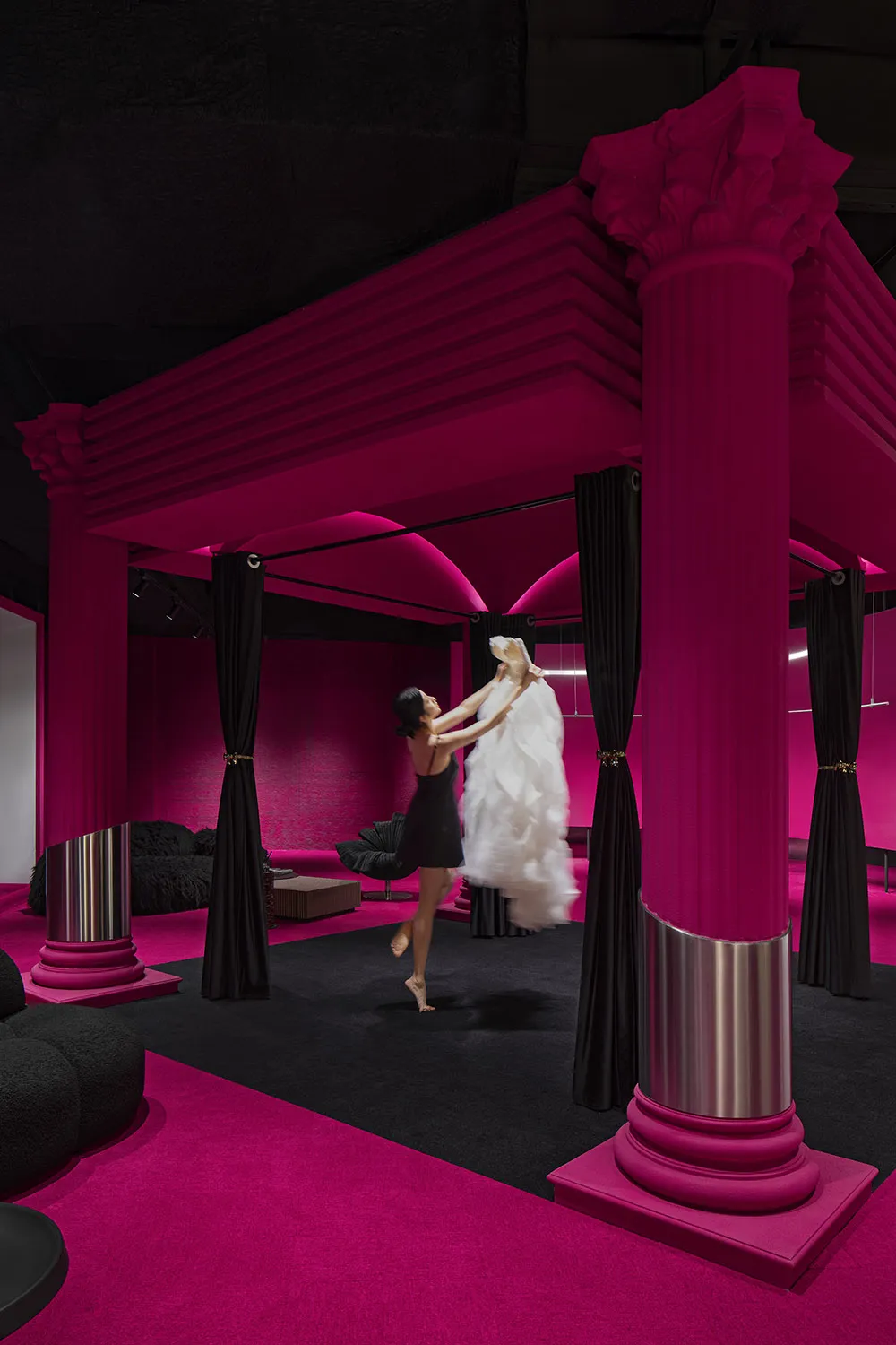 A woman in a black dress holding a white dress on a fashion runway under a pink canopy.