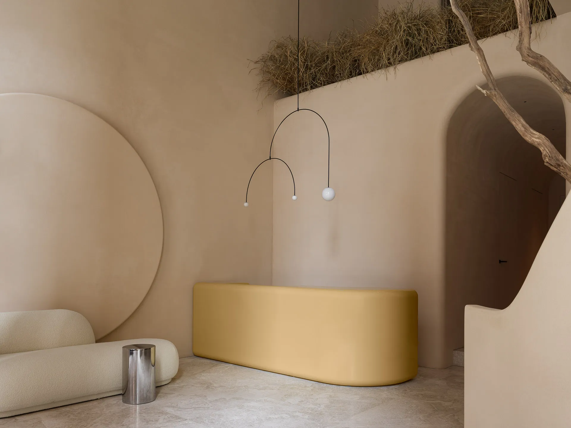 A minimalist interior space featuring a concrete bench and arches, with a metal sculpture hanging overhead and dry grass on t