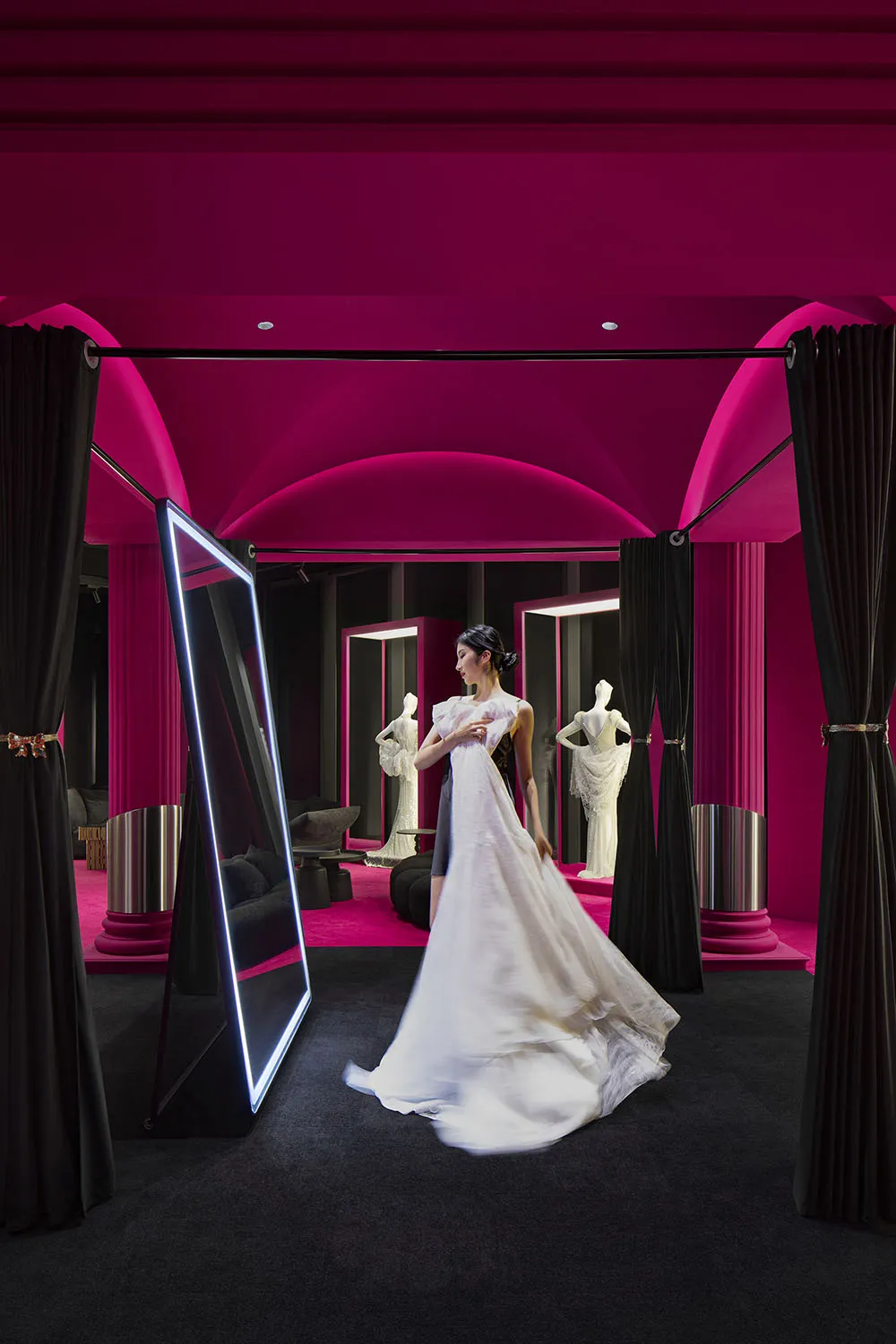 A woman wearing a long white dress in a modern retail space with pink lighting and black curtains.