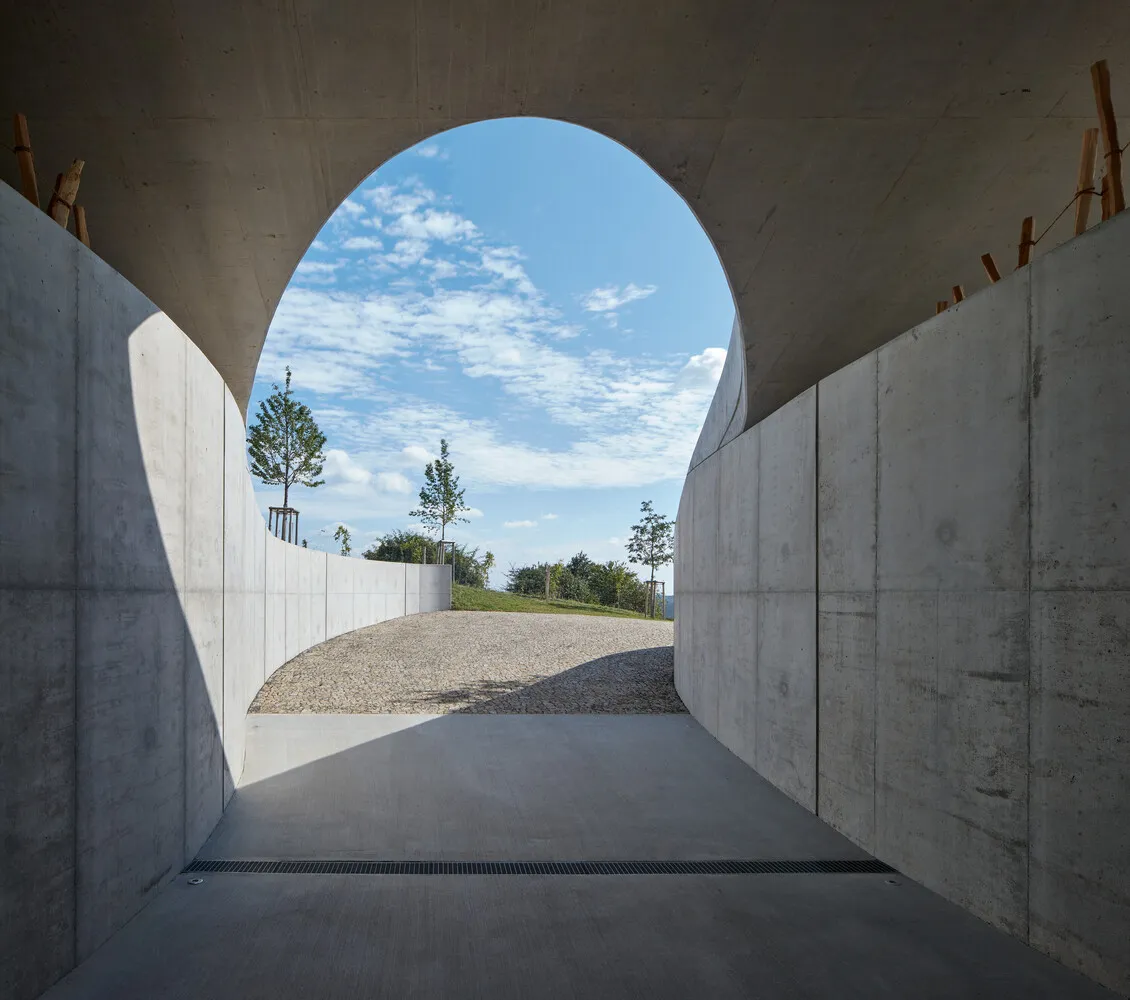 Concrete pathway leading through an archway, surrounded by greenery and light blue sky.
