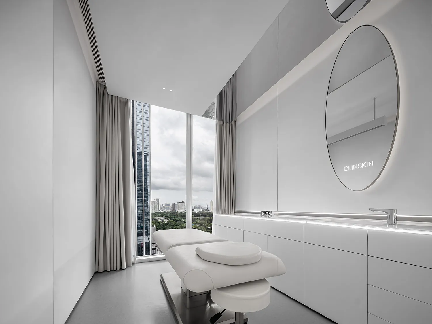 A modern medical office with white walls, a white medical bed, and a large window with a city view.