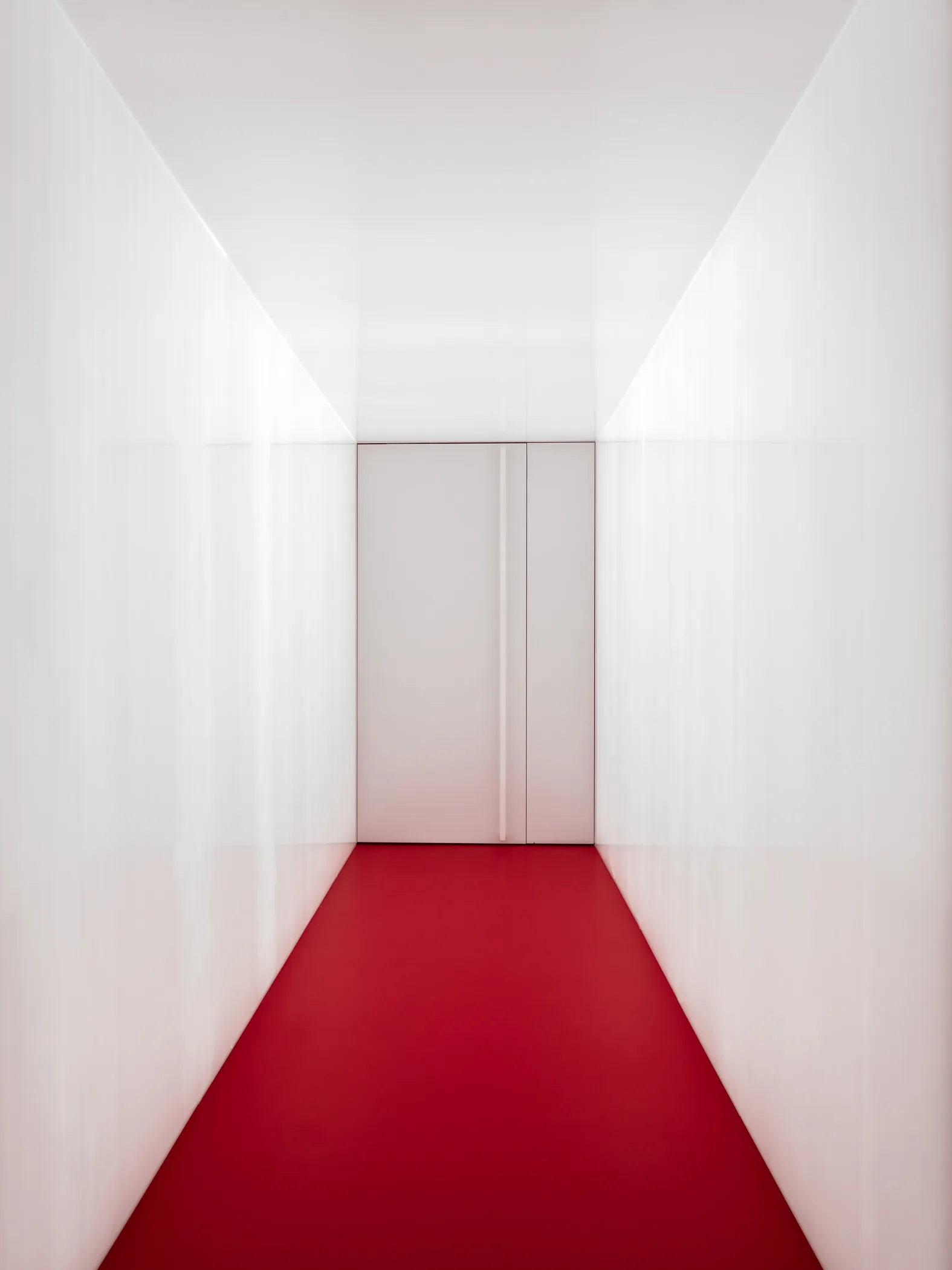 A minimalist corridor with red carpet flooring and white walls, leading to a door.