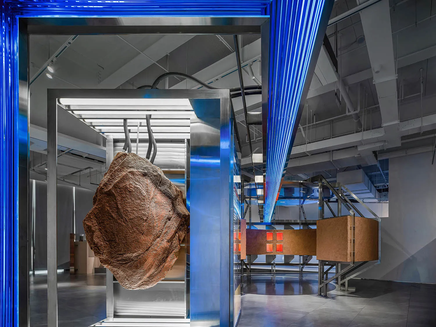 A modern installation art piece featuring a metallic sculpture suspended in a metallic-framed enclosure with blue light beams