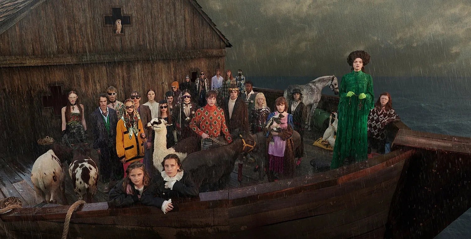 Model wearing a green coat standing on a boat surrounded by animals and people in rain.