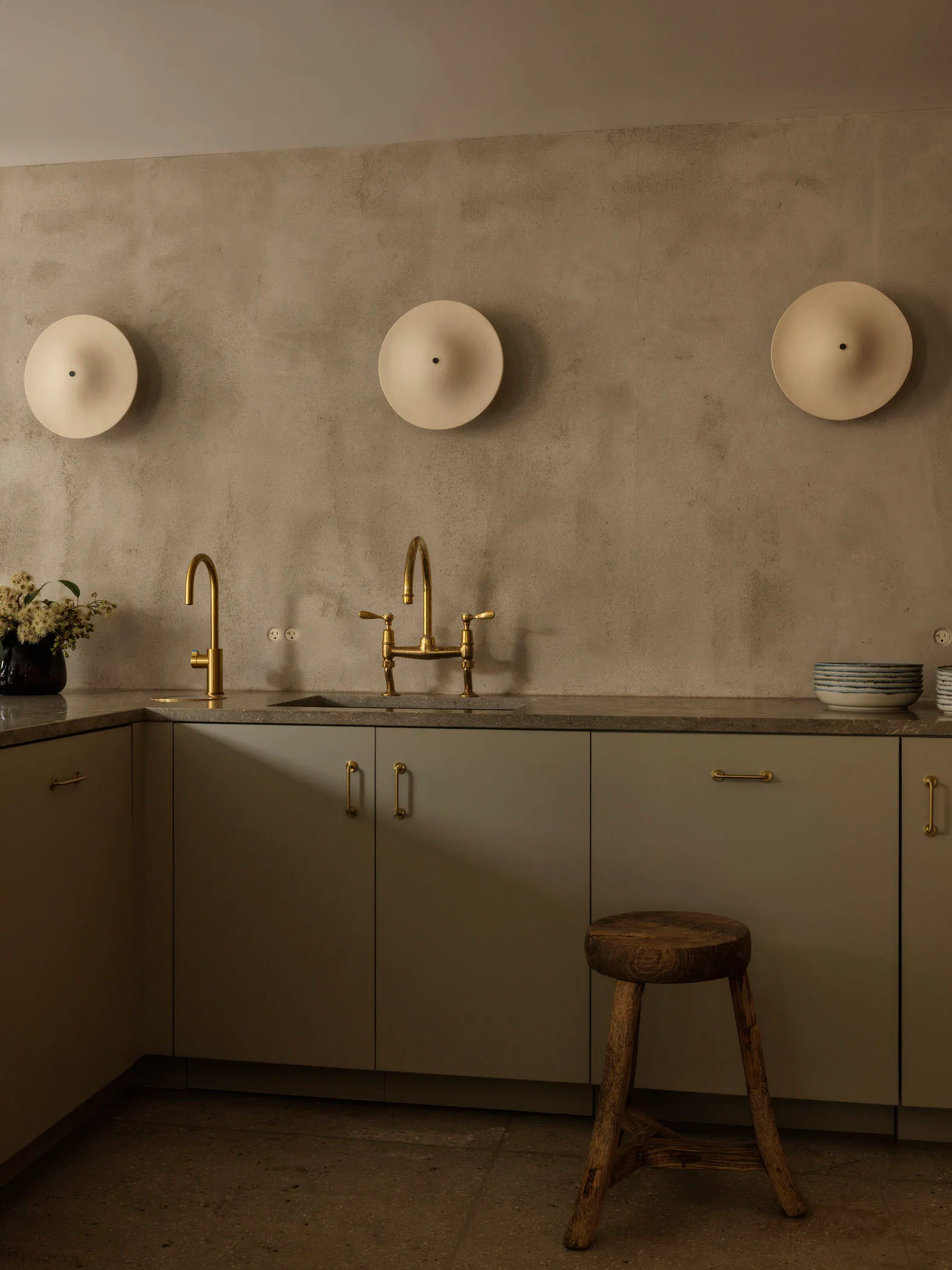 A modern, minimalist residential kitchen featuring concrete countertops and walls, with brass faucets and a stool.