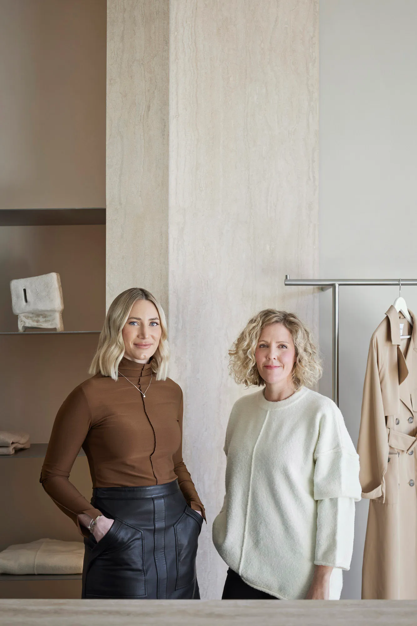 Two women posing in a retail space with neutral-toned clothing.