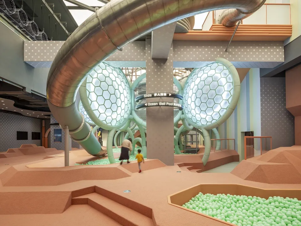 A modern indoor playground featuring metallic slides, a foam pit, and glass enclosures.
