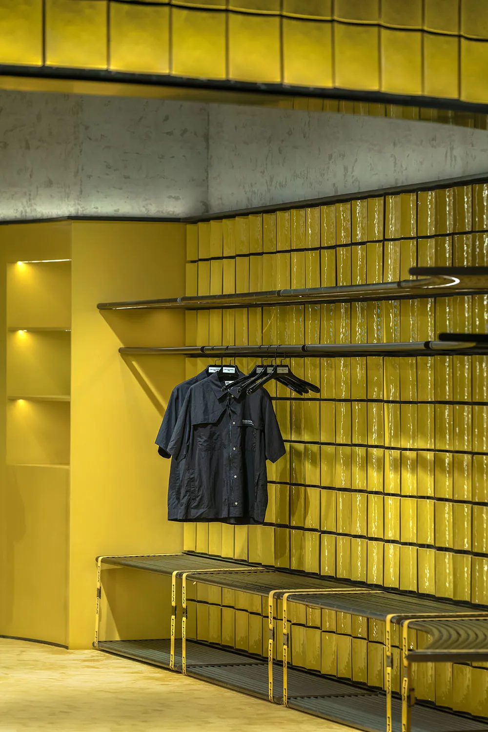 A clothing store with bright yellow ceramic tiles on the walls, concrete flooring, and black metal shelves.