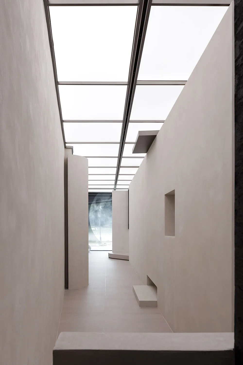 A minimalist corridor with concrete walls and floors, illuminated by a ceiling skylight.