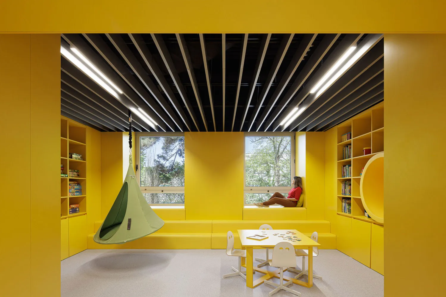 A modern playroom with yellow walls and furniture, featuring a green hanging chair, a white table with chairs, and a window o