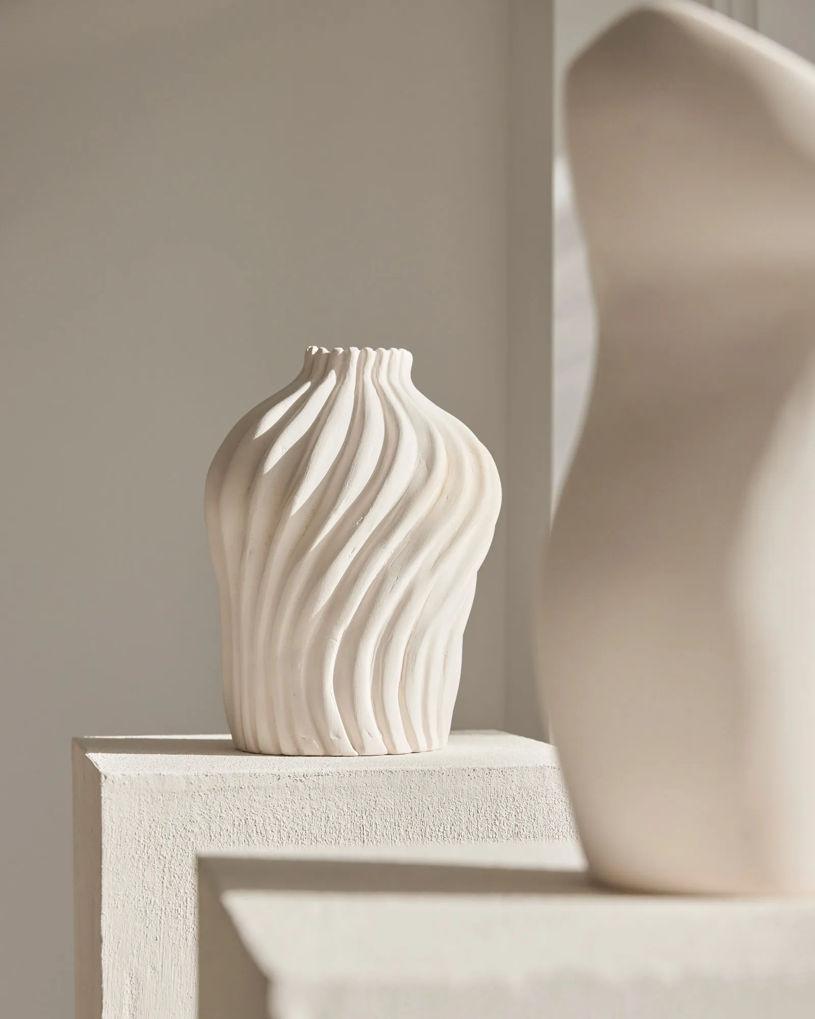 A minimalist, abstract white ceramic vase with wavy lines sits on a white pedestal in a neutral, minimalist setting.
