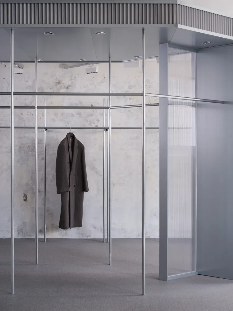 A minimalist showroom featuring a brown coat hanging on a metal frame with glass walls and a textured concrete wall.