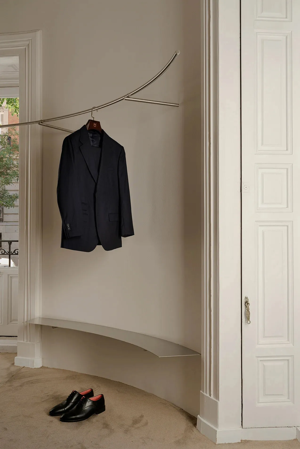 Modern metal and stone wall-mounted clothing hanger with a navy-blue blazer hanging on it, near a pair of black shoes on the