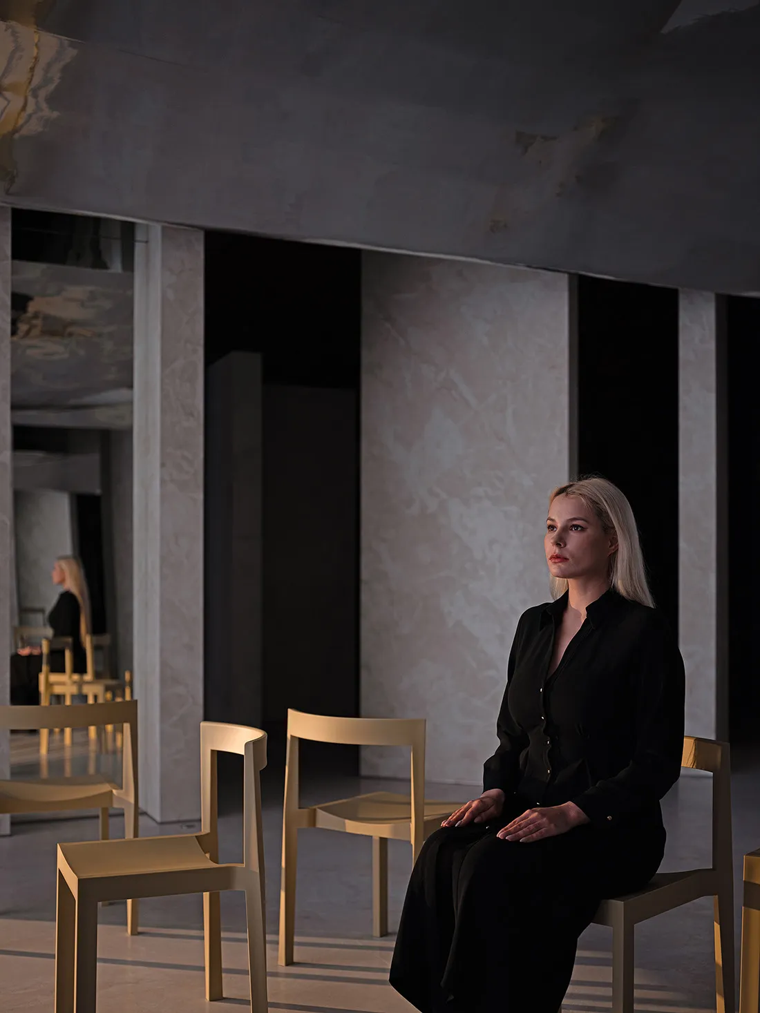 A minimalist interior space with neutral tones, featuring a woman sitting on a chair.