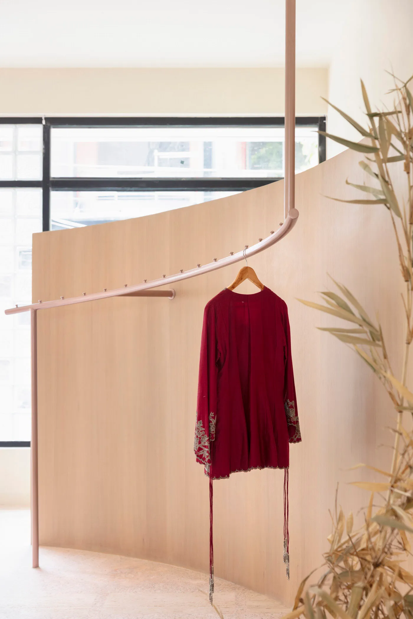 金屬衣架上展示著一件酒紅色刺繡外套,背景為木質展示牆與一片綠葉植物。