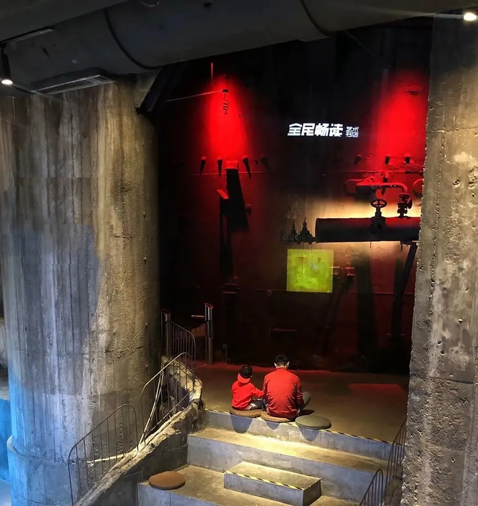 兩位人士坐在博物館展覽中的階梯上,展品包括混凝土管與紅光照亮的畫作。
