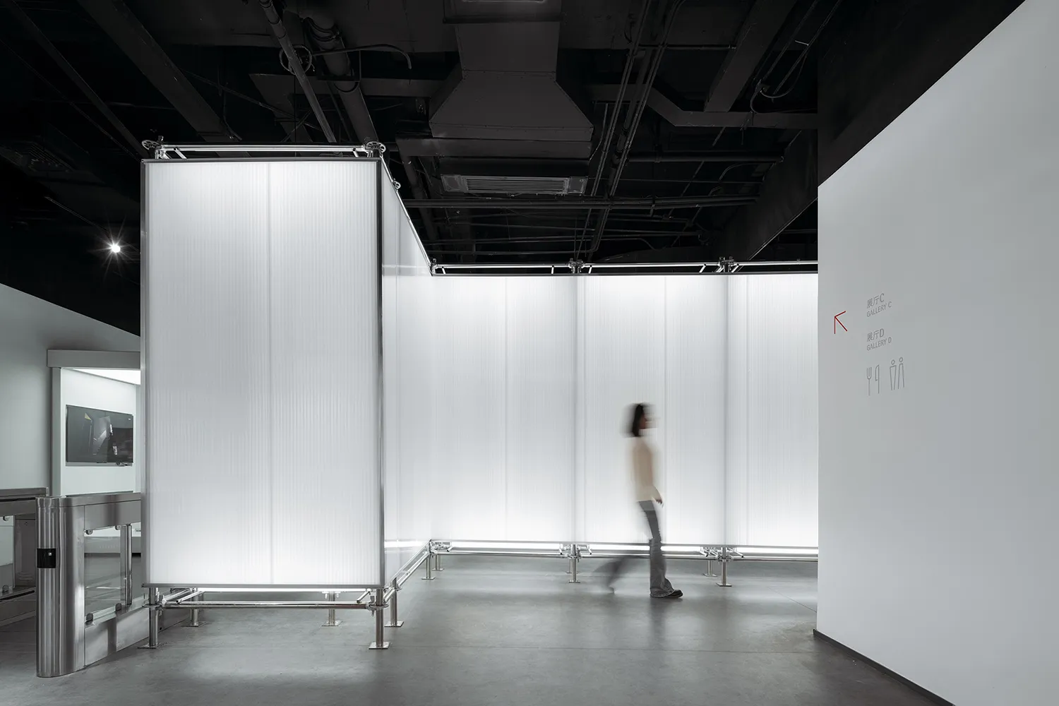 A minimalist exhibition space featuring glass walls and a single blurred person walking through the space.