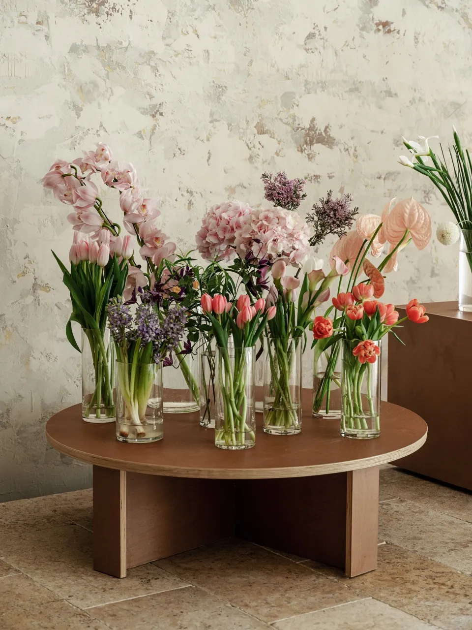 A collection of flowers arranged in clear glass vases on a wooden table against a textured wall. The display includes various