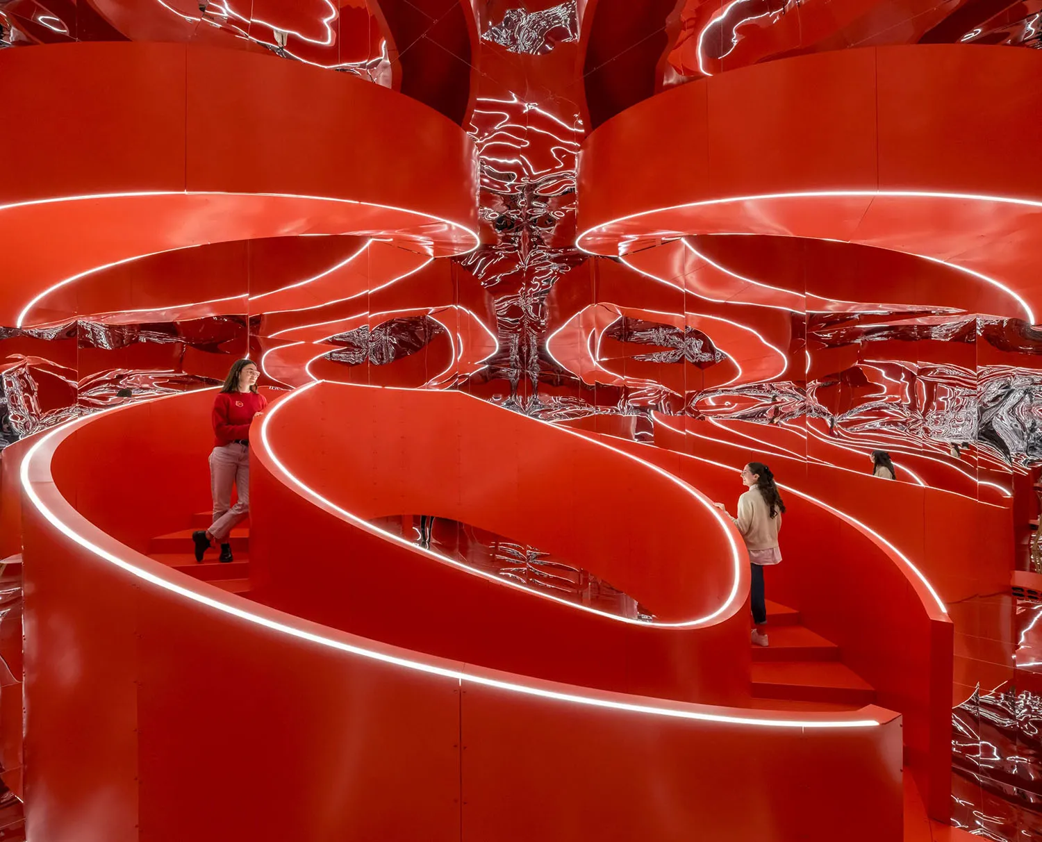 Two people standing on a spiral staircase surrounded by red metallic walls with neon lighting.