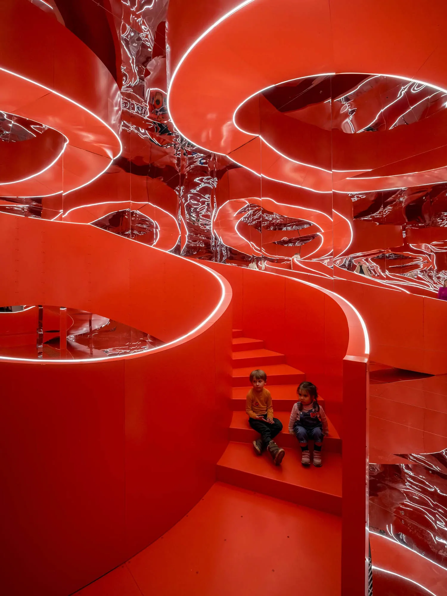 Children sitting on stairs surrounded by red metallic walls with neon lighting and reflective surfaces.