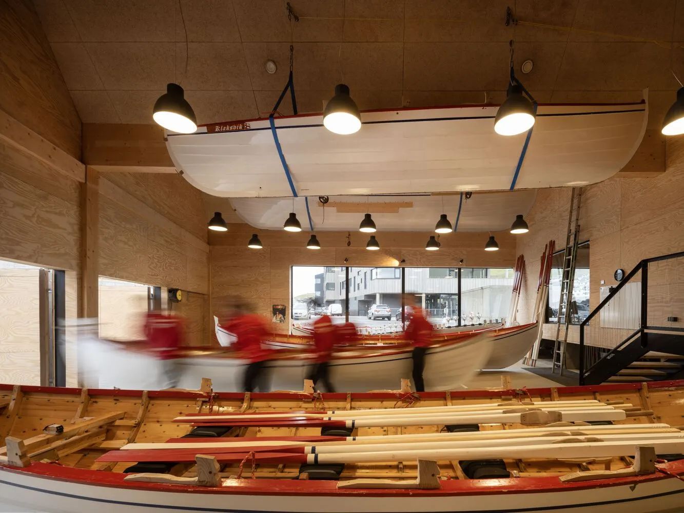 A boatyard workshop with wooden boats and oars on the floor, wooden walls, and hanging lights.