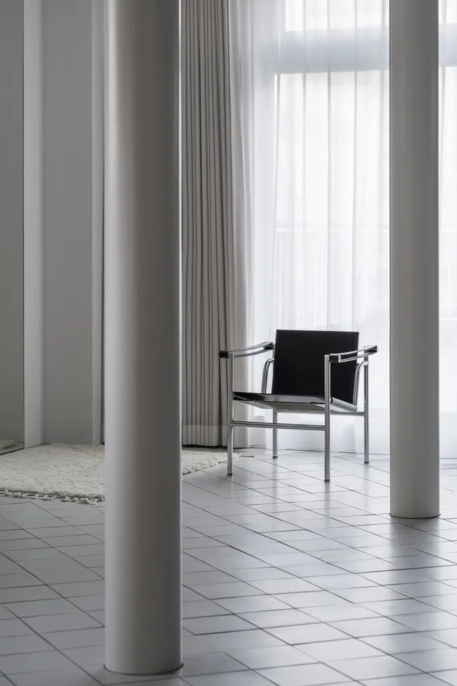 Minimalist design featuring a black metal and leather armchair in a bright, modern room with white flooring and curtains.