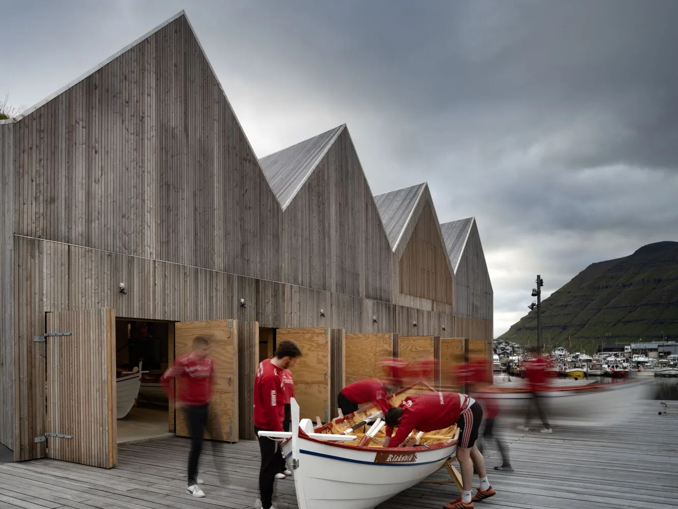 Men working on a boat in front of a modern boat harbor with wooden houses.