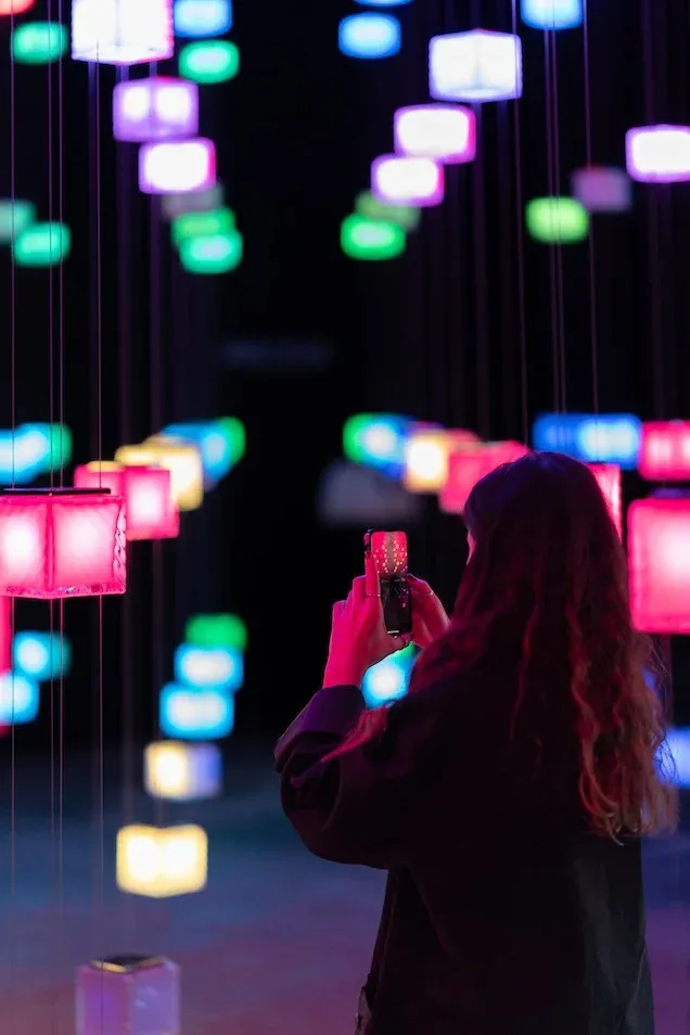 A woman taking a photo of vibrant, hanging LED lights in a dark space.