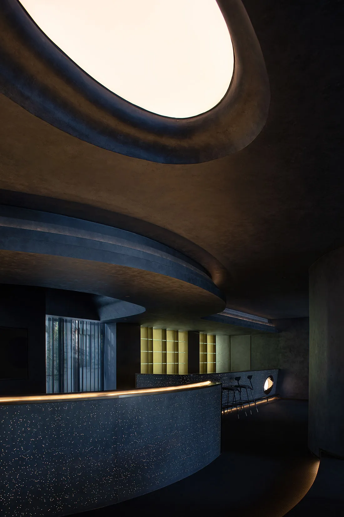 Modern hotel lobby interior featuring a sleek, curved concrete reception desk with a skylight overhead.