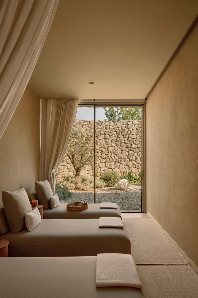 A minimalist bedroom with neutral tones, featuring twin beds, a window with curtains, and a view of a rocky garden.