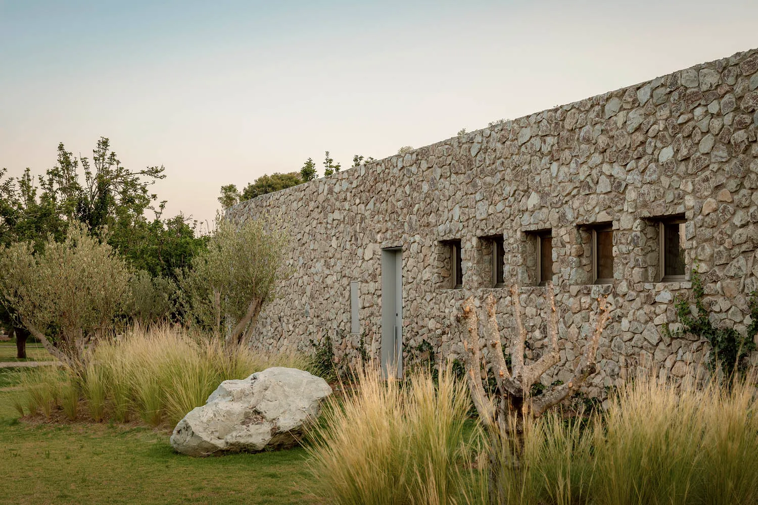 A modern stone building exterior at dusk, surrounded by natural vegetation.