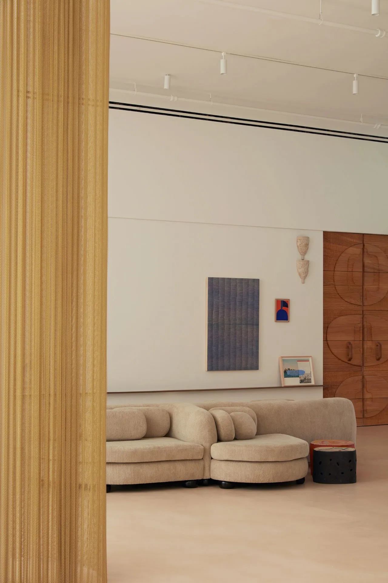 A modern living room with a beige sofa set against a white wall, featuring abstract art and a wooden cabinet. The floor is li